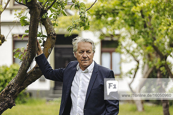 Porträt eines älteren Geschäftsmannes  der an einem Baum in einem ländlichen Garten steht