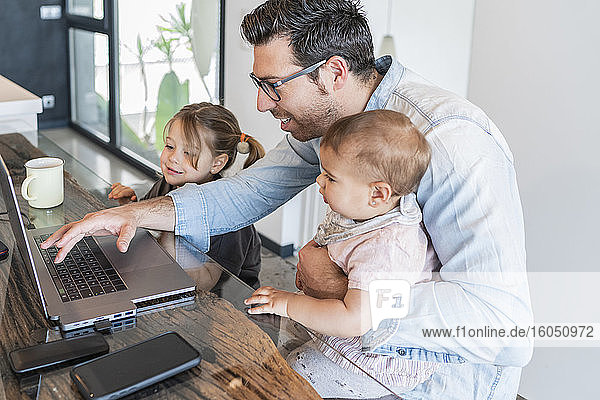 Vater arbeitet über Laptop auf Tisch mit Töchtern an seiner Seite zu Hause