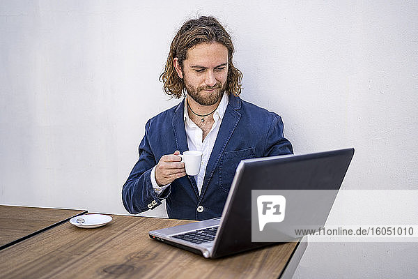 Junger männlicher Unternehmer  der Kaffee trinkt und einen Laptop an der weißen Wand eines Cafés benutzt