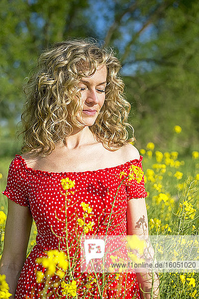 Reife Frau in rotem Kleid mit geschlossenen Augen inmitten von Ölsaatenrapsen stehend