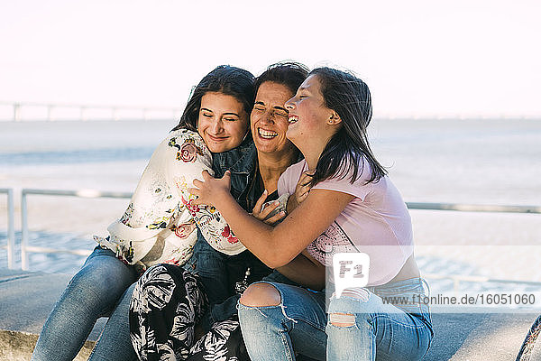 Loving daughters embracing cheerful mother while sitting on retaining wall against sea
