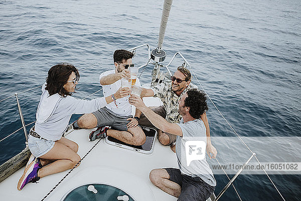 Carefree friends toasting beer glasses while sitting on sailboat in sea