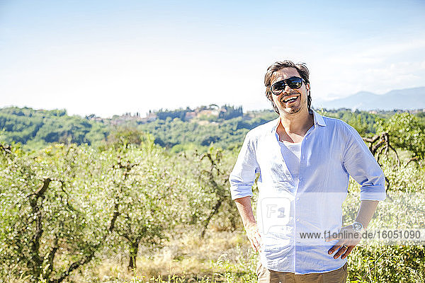 Glücklicher mittelgroßer erwachsener Mann mit Händen auf der Hüfte stehend in einer Olivenplantage gegen den Himmel an einem sonnigen Tag