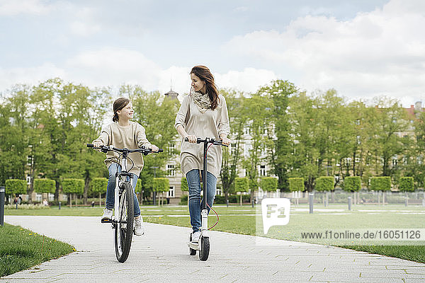 Mädchen fährt Rad  während die Mutter im Stadtpark einen Elektroroller fährt