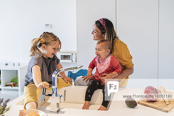 Mutter und fröhliche Töchter spielen mit Wasser in der Küchenspüle zu Hause