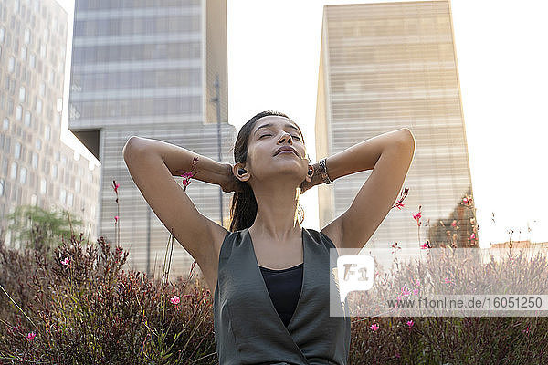 Relaxed young businesswoman listening music through earphones with hands behind head against office building during break
