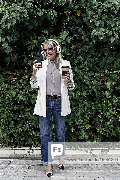 Lächelnde weibliche Fachkraft hört Musik  während sie in der Pause einen Kaffee gegen Pflanzen im Garten genießt
