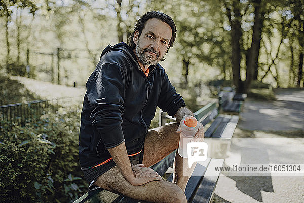 Selbstbewusster reifer Mann  der eine Wasserflasche hält  während er auf einer Bank im Park sitzt
