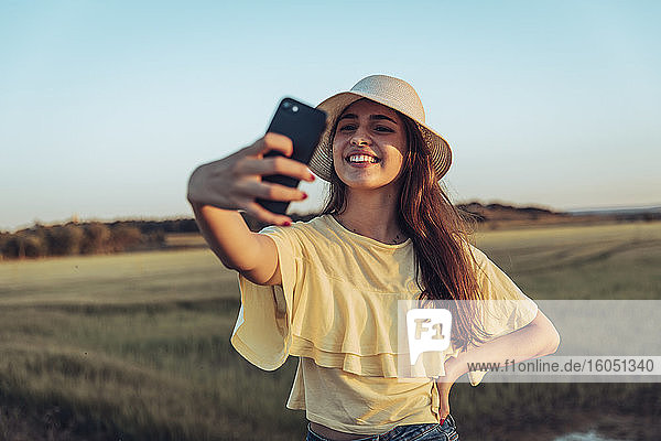 Glückliche junge Frau nimmt Selfie durch Smartphone gegen klaren Himmel bei Sonnenuntergang
