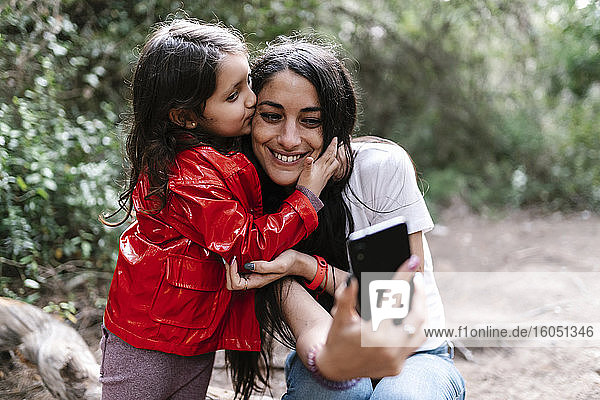 Tochter küsst ihre Mutter  während sie ein Selfie im Wald macht