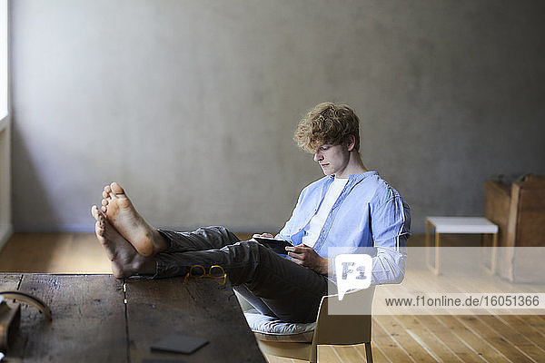 Junger Mann mit Füßen auf dem Tisch  der ein digitales Tablet benutzt