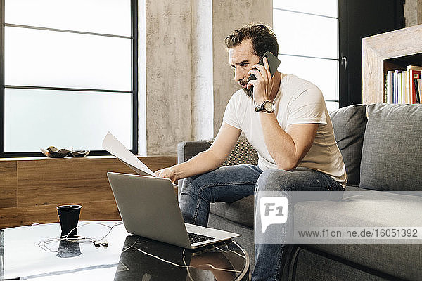 Mature man sitting on couch  using laptop  talking on the phone  holding papers