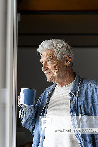 Nachdenklicher älterer Mann lächelt beim Kaffeetrinken am Fenster zu Hause