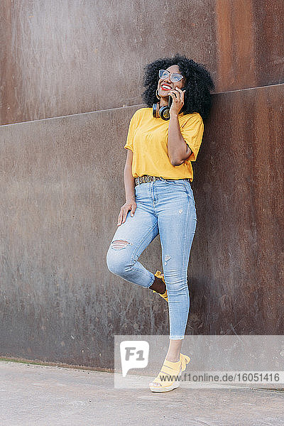 Lächelnde Frau mit Afro-Haar und Brille  die ein Smartphone benutzt und sich an eine Wand lehnt
