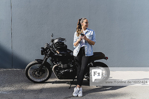 Lächelnde Frau  die auf einem Motorrad sitzt und ein Smartphone benutzt