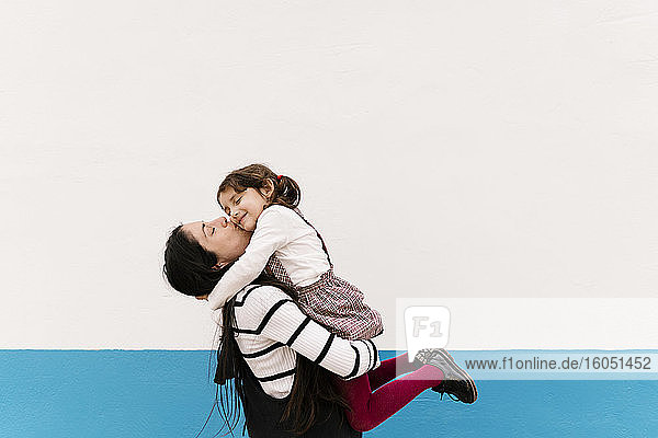 Mother kissing daughter on cheek against wall during sunny day