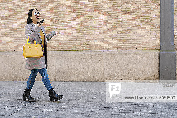 Stilvolle Frau mit Sonnenbrille spricht über Smartphone beim Gehen auf dem Bürgersteig in der Stadt