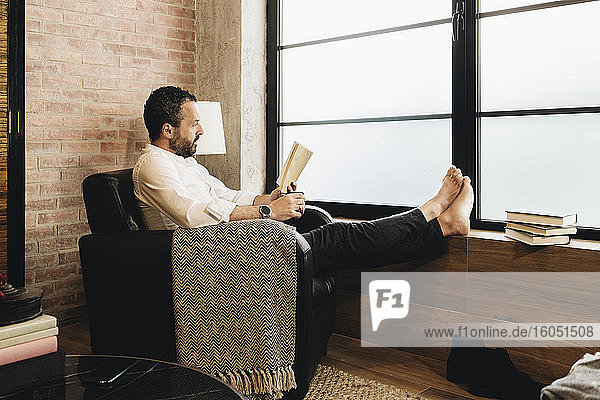 Mature man sitting in armchair by window  reading book