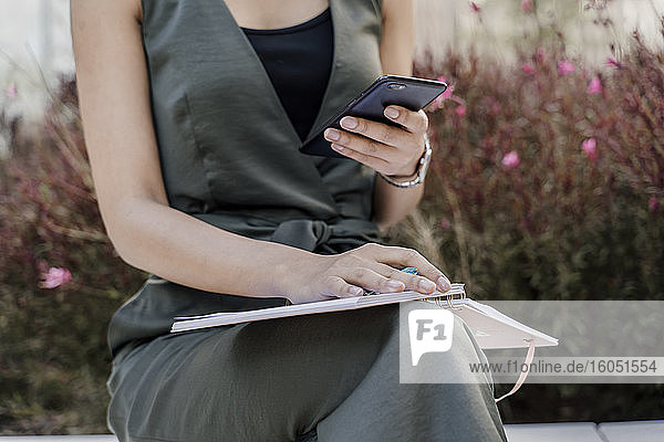 Nahaufnahme einer weiblichen Fachkraft mit Notizblock  die ihr Smartphone benutzt  während sie vor Pflanzen sitzt