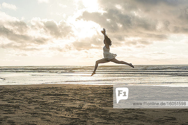 Glückliche junge Frau mit erhobenen Armen springt am Strand gegen bewölkten Himmel bei Sonnenuntergang