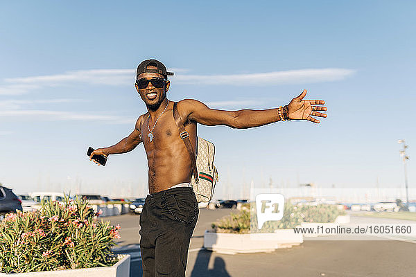 Shirtless young man wearing sunglasses with arms outstretched standing on road in city