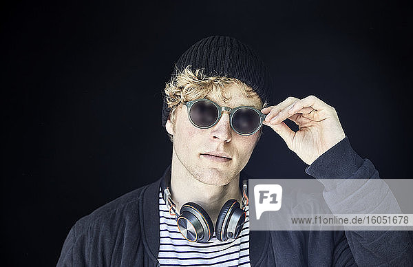 Porträt eines jungen Mannes mit Kopfhörern  Mütze und Sonnenbrille vor schwarzem Hintergrund