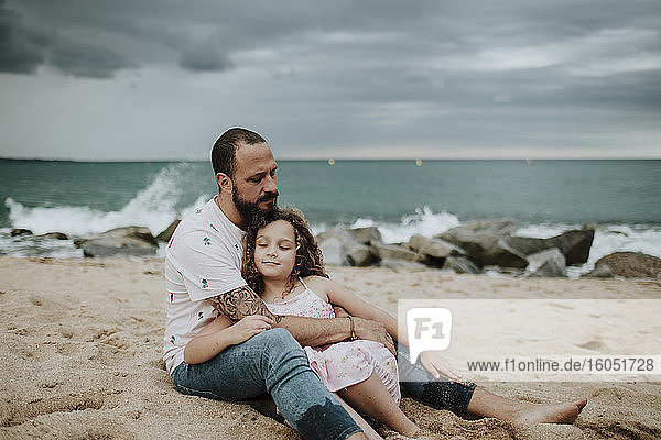 Vater umarmt Tochter am Strand sitzend