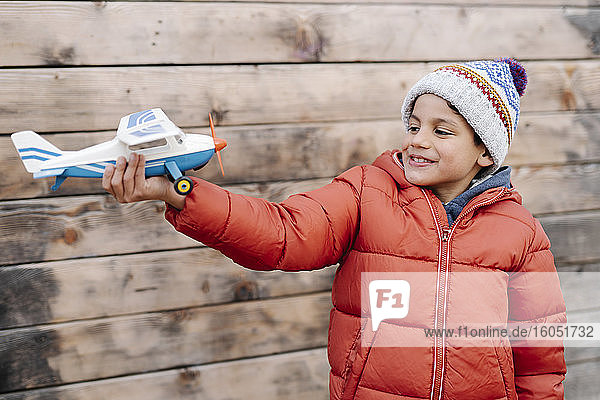 Lächelnder Junge in warmer Kleidung  der ein Modellflugzeug hält und an der Wand steht