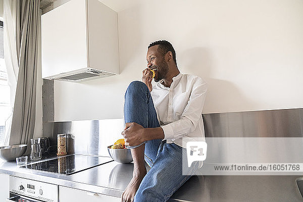 Lachender Mann  der zu Hause auf dem Küchentisch sitzt und einen Apfel isst