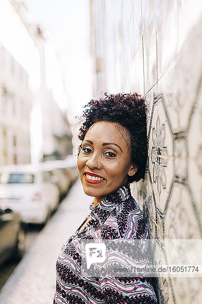 Lächelnde stilvolle Frau lehnt an einer gefliesten Wand in der Stadt