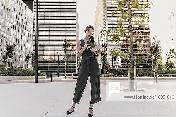 Eine Unternehmerin hält einen Notizblock und benutzt ein Smartphone  während sie vor einem modernen Gebäude in der Stadt steht