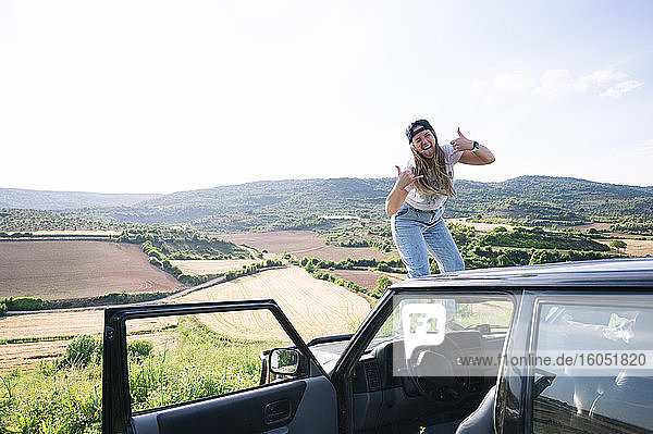 Cheerful woman sticking out tongue while showing hand sign on vehicle hood against sky