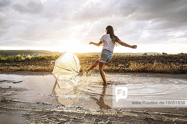 Mädchen spritzt Wasser über Regenschirm auf Pfütze bei Sonnenuntergang