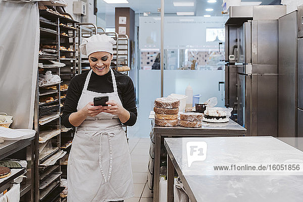 Glückliche Bäckerin  die ihr Smartphone in einer Großküche einer Bäckerei benutzt