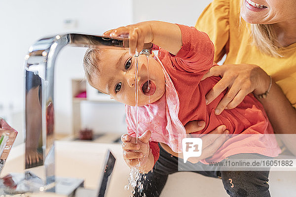 Nahaufnahme einer Mutter  die ein süßes kleines Mädchen hält  das Wasser aus dem Wasserhahn in der Küchenspüle trinkt