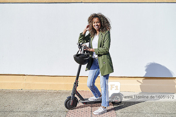 Glückliche Frau steht mit elektrischem Tretroller auf dem Bürgersteig an einem sonnigen Tag