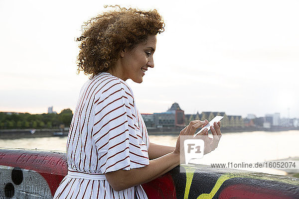 Lächelnde Frau mit lockigem Haar  die ein Smartphone benutzt  während sie an einem Geländer steht und den Himmel bei Sonnenuntergang betrachtet