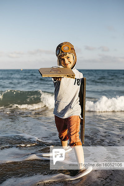 Boy with aviator's cap holding cardboard wings while standing at beach