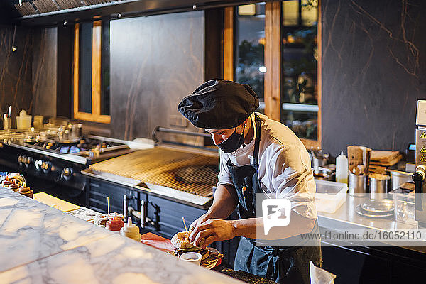 Koch mit Gesichtsschutzmaske bei der Zubereitung von Burgern in der Restaurantküche