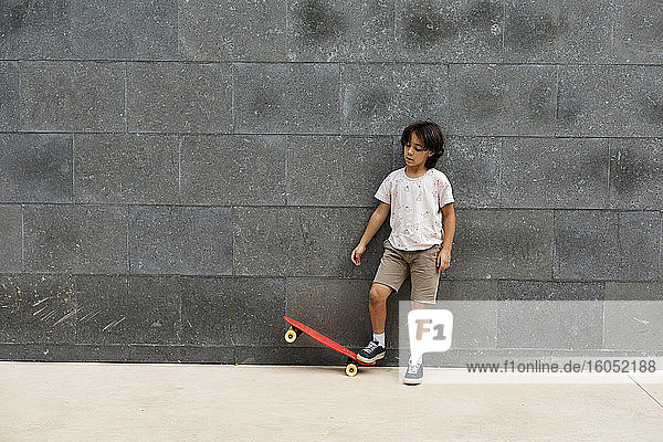 Boy playing with skateboard while standing on footpath against wall