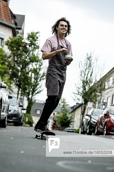 Lächelnder männlicher Barista  der eine Kaffeetasse hält  während er auf einer Straße in der Stadt Skateboard fährt
