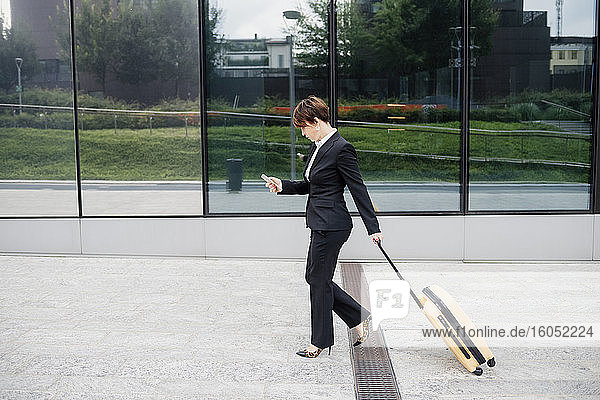 Unternehmerin  die ein Mobiltelefon benutzt  während sie mit einem Koffer auf dem Fußweg geht