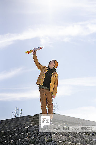 Boy holding rocket toy while standing on steps against sky
