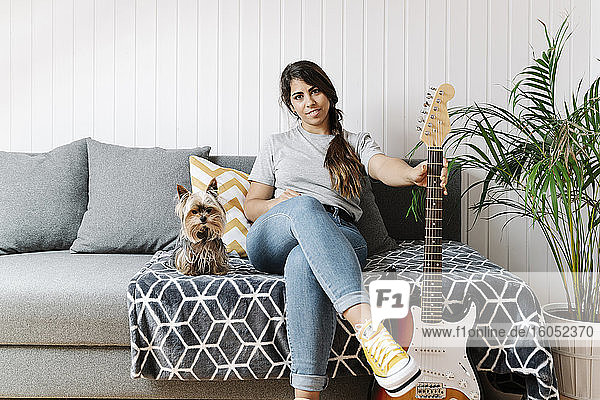 Selbstbewusste Frau  die eine E-Gitarre in der Hand hält  während sie mit ihrem Yorkshire Terrier auf dem Sofa zu Hause sitzt