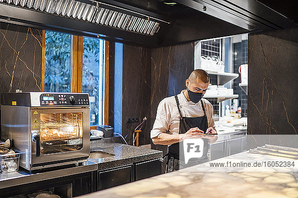 Chef wearing protective face mask using smartphone in restaurant kitchen