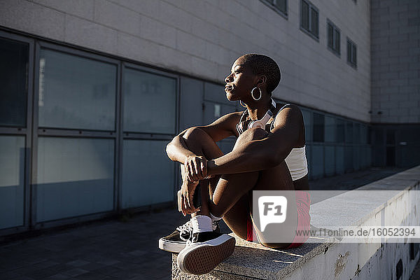 Nachdenkliche junge Frau mit rasiertem Kopf  die auf einer Stützmauer vor einem Gebäude sitzt