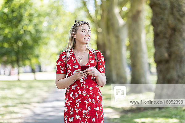 Nachdenkliche Frau  die ihr Smartphone in der Hand hält  während sie auf dem Fußweg in einem öffentlichen Park spazieren geht