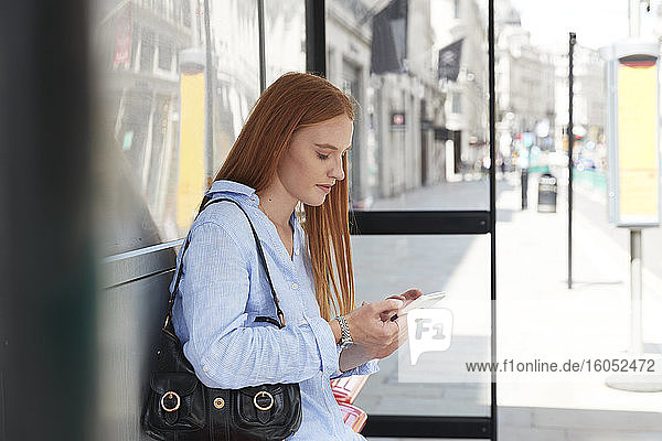 Schöne rothaarige Frau  die ein Mobiltelefon benutzt  während sie an einer Bushaltestelle in der Stadt wartet