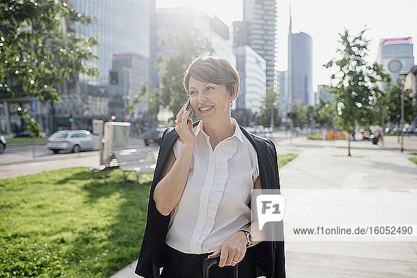 Smiling businesswoman talking over smart phone looking away while standing on sidewalk in city
