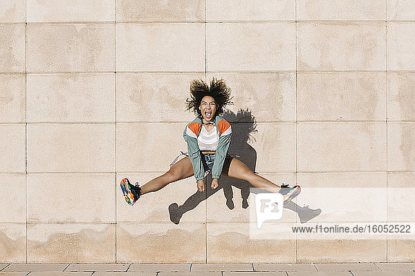 Excited young woman screaming while jumping against wall during sunny day